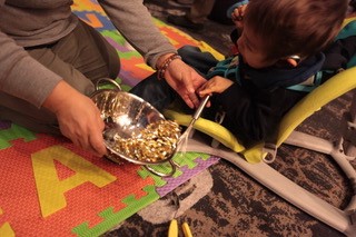 A young boy sitting in a chair holding a whisk. An adult kneels beside him, holding a metal bowl with gold beads inside