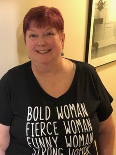Portrait of a smiling woman with short red hair in a black shirt with the words, “Bold Woman, Fierce Woman, Funny Woman, Strong Woman” in white lettering on the front.