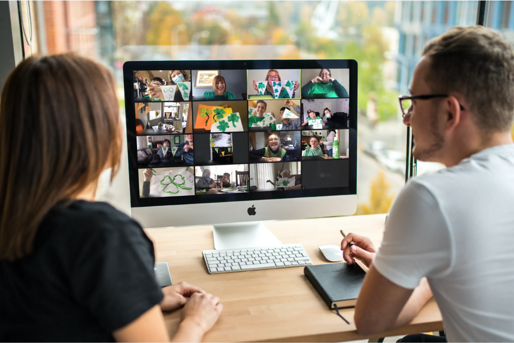 Picture of a computer monitor with a collage of 16 photos of people holding up their St. Patrick’s Day green shamrock paintings for their Zoom activity. A woman and a man are in the foreground sitting at a desk and looking at the computer monitor.