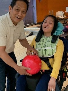 A smiling man with a white shirt standing beside his daughter, who is wearing a yellow shirt and green apron and is in a wheelchair. They are both holding a bright pink bowling ball.