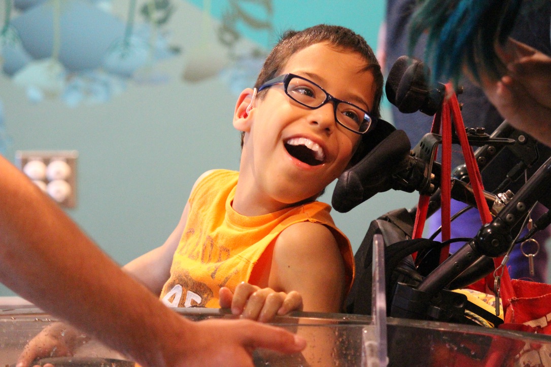 A boy in a yellow shirt sitting in his wheelchair laughing. He is looking to his left at someone