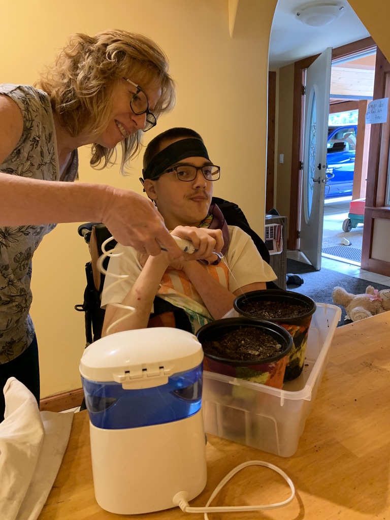 A smiling woman is leaning over a young man in a wheelchair at a table. She is helping him to hold a water pick to water his potted plants.
