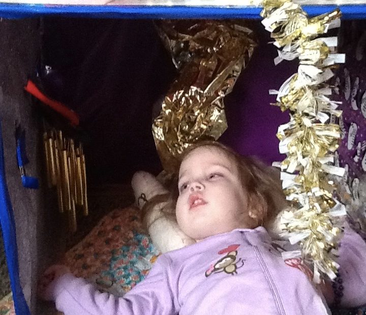 A young girl in a pink outfit I lying down and looking up at gold chimes, gold paper and gold garland hanging down from above her.