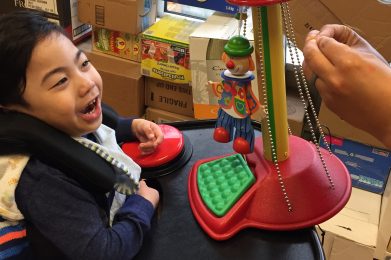 A young boy with dark hair in wheelchair using a large red switch. He is laughing and looking at a wooden clown hanging in front of him, while an adult signs ‘more’ in front of him.