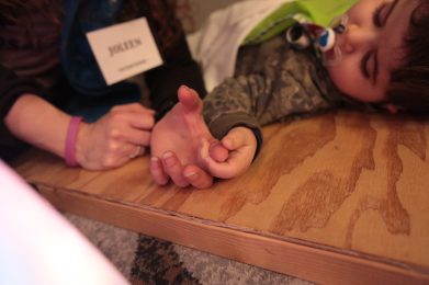 A closeup of a child’s hands holding the pointer finger of an adult hand. The child is laying on his back with a soother in his mouth.