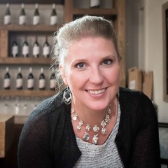 Portrait of a smiling woman with blonde hair tied back in a black and grey shirt with a beaded necklace and earrings.