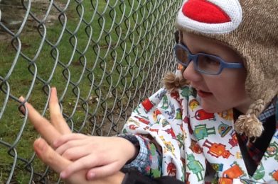 A young boy with blue glasses and a brown hat with a monkey face sitting in front of a chain-link fence. His hand is on top of an adult hand on the fence.