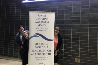 A white with blue lettering banner with ‘June is Deafblind Awareness Month, Make a Wave From Coast to Coast’. Two woman are peeking out from behind the banner to the left and one woman to the right.