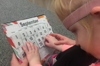 A young girl looking at and holding a monthly calendar of September. An adult hand is supporting her right hand.