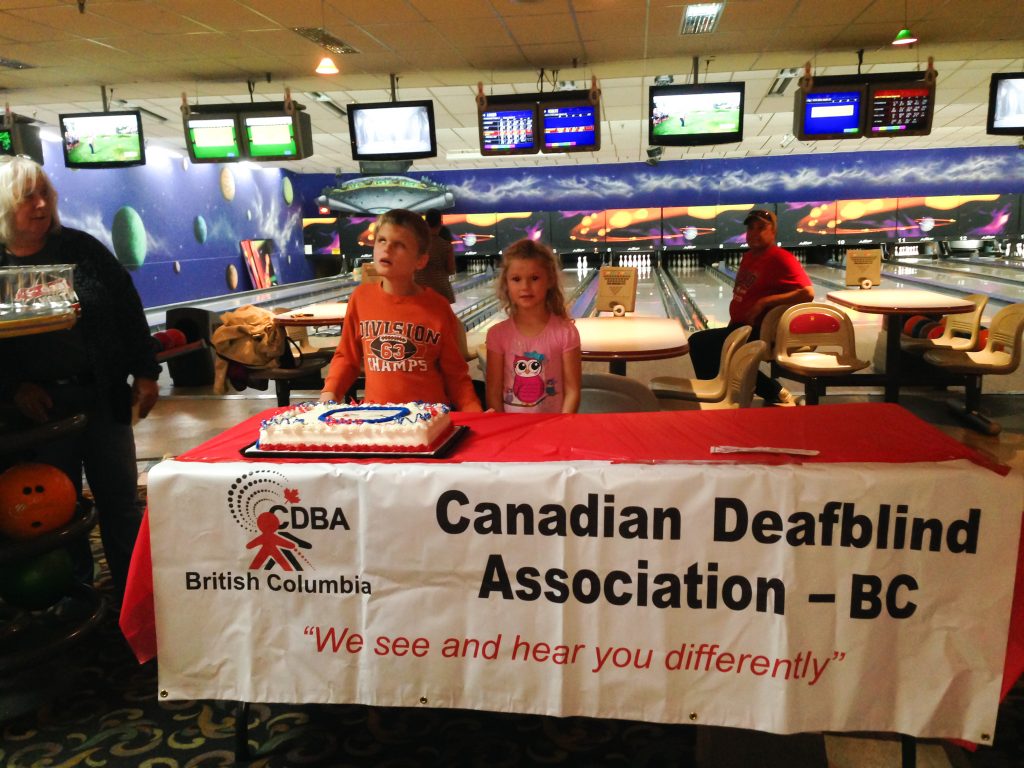 A boy with short blonde hair wearing an orange long-sleeved shirt is standing behind a rectangular table with a red tablecloth and a white banner across the front with the words ‘Canadian Deafblind Association of BC’. To the boy’s right is a girl with long blonde hair wearing a pink t-shirt with a pink owl on the front. On the table in front of them is a rectangular cake with white icing and blue and red piped icing.