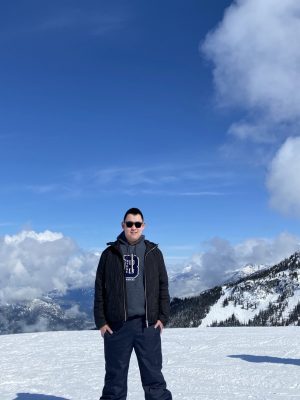 A young man with short dark hair and sunglasses is wearing a dark snow suit and standing on a field of snow with clouds and mountains in the background.