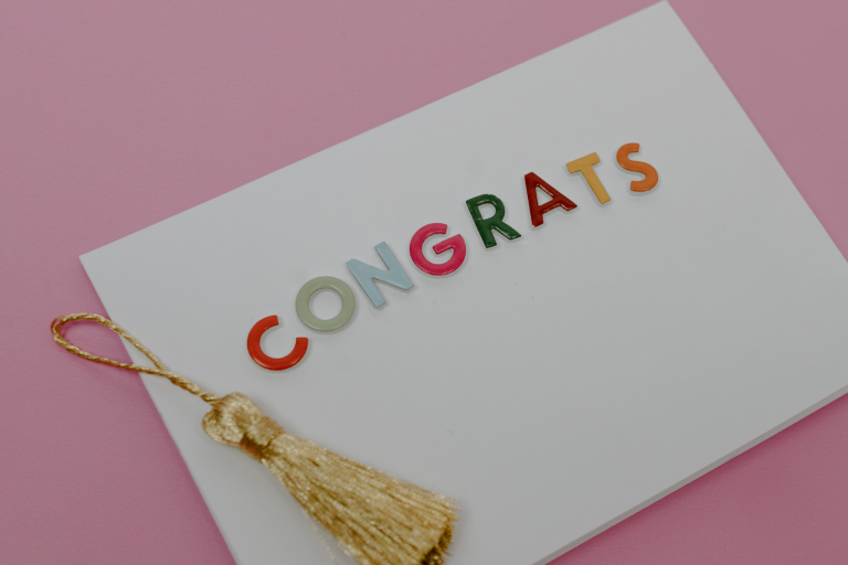 A photo of a white card with ‘Congrats’ on the front in coloured letters. A gold tassel lays on the left side of the card.