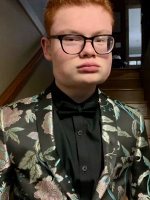 A young man with red hair and black glasses wearing a black suit jacket with shiny green leaves and pink flowers and a black dress shirt beneath poses for the camera.