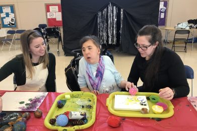 A girl wearing a blue shirt and scarf is sitting in her wheelchair. On her right is a woman with long brown hair wearing a black hair band, black shirt and white apron. She is smiling while watching the young girl paint. On the girl’s left is a woman with black hair in a pony tail wearing a black shirt. Her hand is under the girl’s hand while she holds a pink ball dipped in yellow paint. They are using the ball to paint a white board that is on a yellow tray. A large black tent with silver streamers in the doorway is behind them.