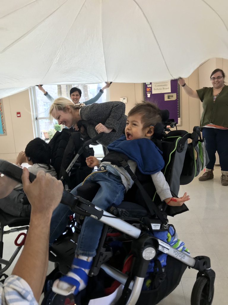 A young boy with short brown hair wearing a grey sweatshirt and jeans is in his wheelchair, looking up and laughing. Above him is a white parachute suspended in the air.