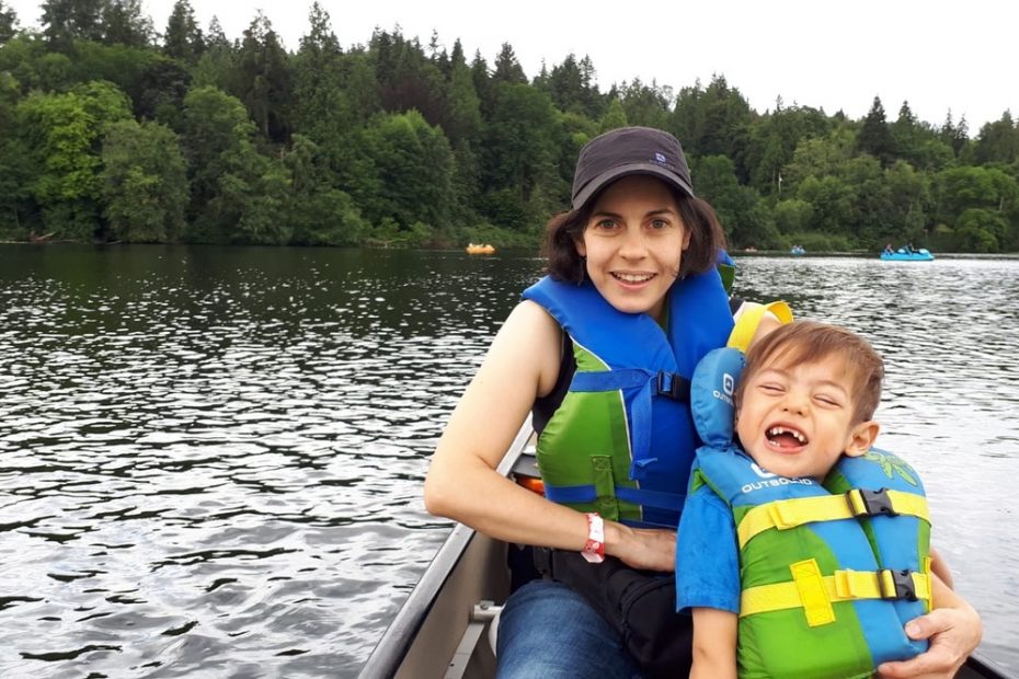 A smiling woman with dark hair wearing a black hat and blue and green lifejacket is sitting in a canoe on a lake. In front of her, leaning against her, is a laughing young boy with brown hair and a blue and green lifejacket with yellow straps.