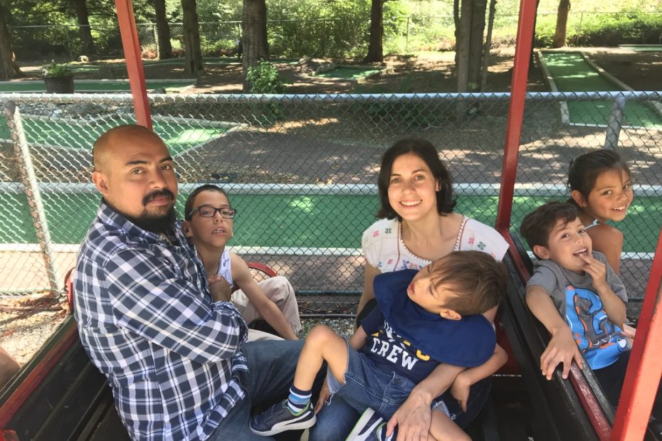 A balding man with a mustache and beard, wearing a blue and white checkered shirt is sitting in the forefront beside a young boy with dark hair and glasses. They are sitting on a park train car across from a woman with dark hair and a white shirt who is supporting a young boy with brown hair and a blue shirt on her lap. Beside them on another train car is a young girl with dark hair and a young boy with dark hair and a grey and blue shirt. All six individuals are looking up and smiling for the photo.