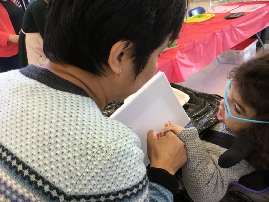 A woman with dark hair and light blue sweater is looking at a young girl in a wheel chair who has blue glasses and a grey sweater. They are holding hands and painting a picture together, using the hand-under-hand technique.