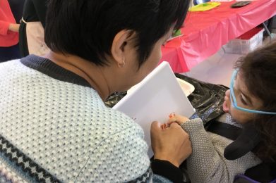 A woman with dark hair and light blue sweater is looking at a young girl in a wheel chair who has blue glasses and a grey sweater. They are holding hands and painting a picture together, using the hand-under-hand technique.