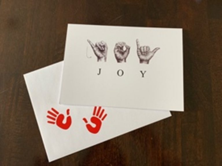 A greeting card on top of an envelope with two red open hand imprints in the bottom left corner. Horizontally across the center of the card are the sketched ASL letters to spell ‘JOY’. Below the sketched hands are the corresponding individual letters.