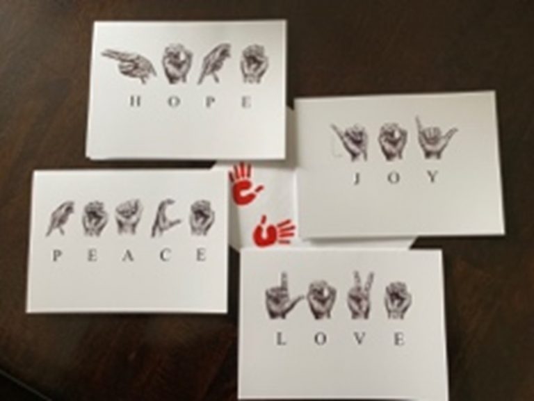 Photo of 4 Greeting cards around 1 envelope with two red hand prints on it. One card has the sketched ASL letters to spell ‘HOPE’, one card has the sketched ASL letters to spell ‘LOVE’, one to spell PEACE’ and one to spell ‘JOY’. The corresponding letters are below each sketched hand.