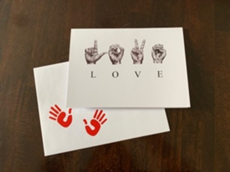 A greeting card on top of an envelope with two red open hand imprints in the bottom left corner. Horizontally across the center of the card are the sketched ASL letters to spell ‘LOVE’. Below the sketched hands are the corresponding individual letters.