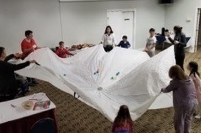A group of women and children standing in a room in a circle around a white parachute. There are several small coloured balls on the parachute and the group is holding the parachute up in the air.