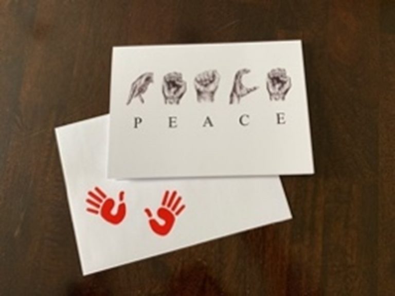 A greeting card on top of an envelope with two red open hand imprints in the bottom left corner. Horizontally across the center of the card are the sketched ASL letters to spell ‘PEACE. Below the sketched hands are the corresponding individual letters.