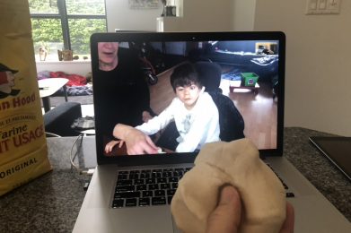 A laptop shows a picture on its screen of a boy with short dark hair in a long-sleeved white shirt looking forward. To his left is his mother with a long-sleeved black shirt pointing her hand in front of him and also looking forward. In front of the laptop is a hand holding a ball of dough, holding it up to the computer to show the boy and his mother. To the left of the computer is a yellow bag of Robin Hood flour.