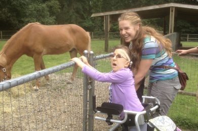 A young girl in a walker wearing a mauve shirt and a bone conduction hearing device is with a laughing woman with long hair and a green, blue and purple striped shirt. They are looking to the left for the photo and standing at a chain-link fence in front of a brown horse.