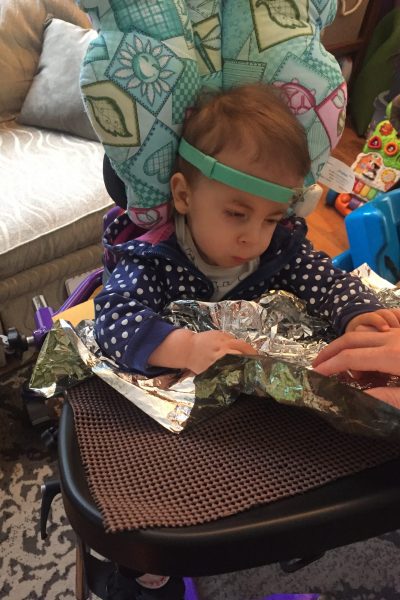 Young girl in blue shirt with white polka dots in a wheelchair wearing a green bone conduction hearing device and playing with shiny crinkle paper. She has her hand on top of an adult’s hand, using Hand-under-hand exploration.