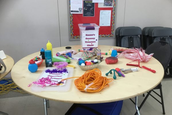 A photo of a table with a clear bin on it labeled “Emma’s Sensory Treasure Chest”. On the table are many colourful sensory items, such as a large roll of orange twine, a dish of coloured pom-poms, pink ribbon, purple feathers, silver and white measuring spoons and a yellow squeeze bottle.