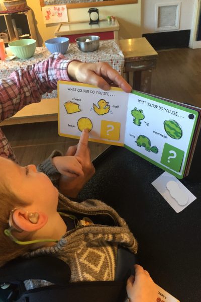 A young boy with hearing aids is in his wheelchair looking at a book with pictures of yellow and green objects. The boy has his hand resting on an adult’s hand, while they point out the objects, using hand-under-hand technique.