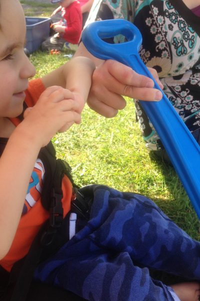 A young boy in blue pants and an orange shirt clasping his hands in front of his face. He is looking at a blue wagon handle that is held by an adult.