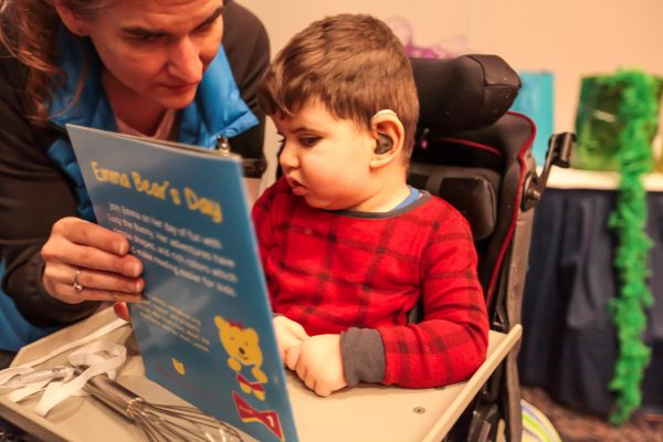 A young boy with short dark hair and hearing aids wearing a red and black checked shirt is sitting in his wheelchair. He is looking at a blue book titled, “Emma Bear’s Day”. To his right is a woman who is holding the book and looking at the book with him. On his wheelchair tray behind the book is a silver whisk.
