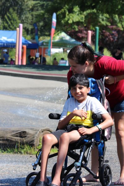 A smiling boy wearing a white and yellow t-shirt and shorts is in a wheelchair at the water park. Leaning over him from behind is a woman with long brown hair in a pony tail wearing a red t-shirt and denim shorts. To their right is a log lying on a strip of grass with a spray of water shooting up from the paved surface.