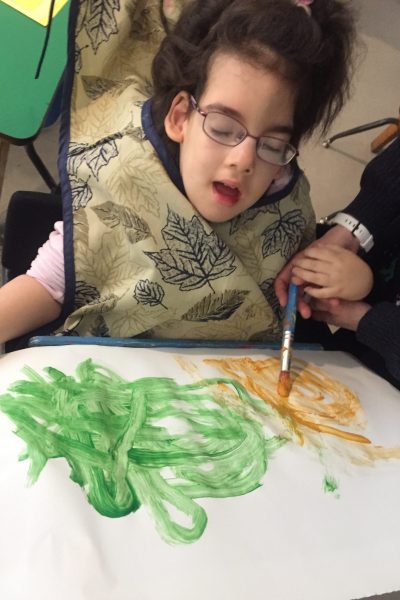 A young girl with dark wavy long hair wearing a bib with sketches of maple leaves on it is painting an orange and green picture. An adult on her left is supporting her left hand as they use hand-under-hand technique to paint with a blue paint brush.