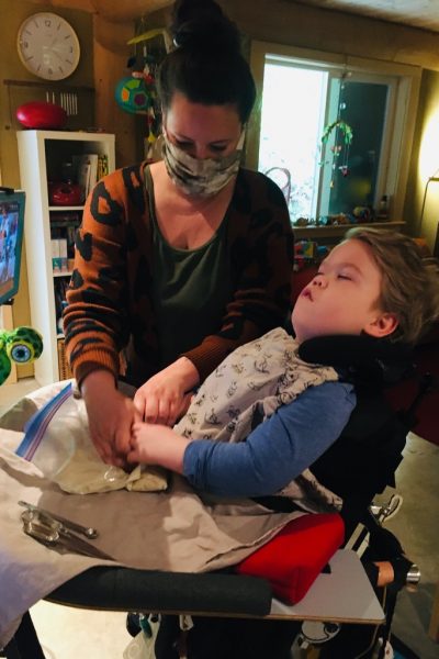 A young boy in a wheelchair is wearing a blue shirt and white bib. He is looking at his computer mounted in front of him. His Intervenor has dark hair tied in a bun and is wearing a mask over her mouth and a green shirt with an orange and black patterned cardigan. The Intervenor is supporting the boy’s hands while they squish white dough in a Ziploc bag.