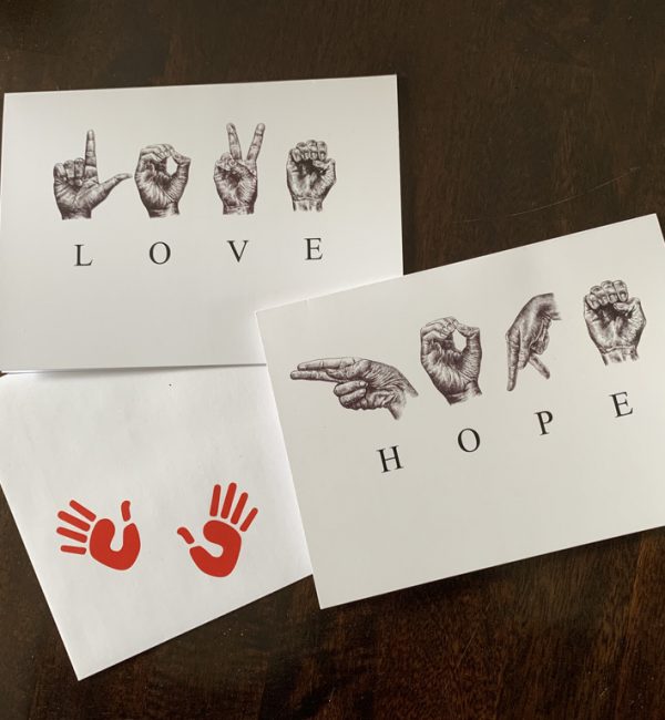Photo of 2 Greeting cards and 1 envelope with two red hand prints on it. One card has the sketched ASL letters to spell ‘HOPE’ and one card has the sketched ASL letters to spell ‘LOVE’.