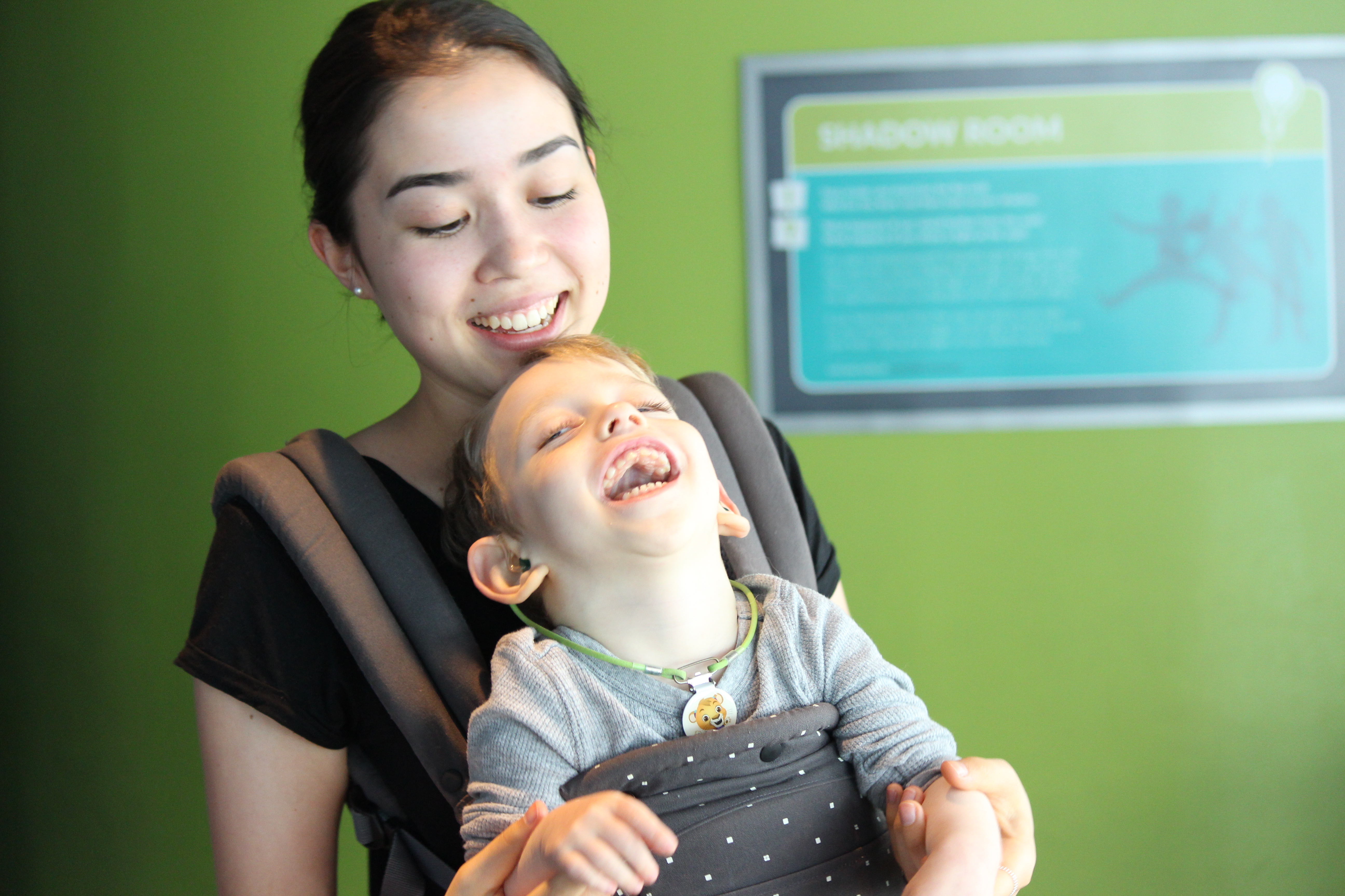 A smiling woman wearing a front pouch. A young boy is in the pouch, laughing