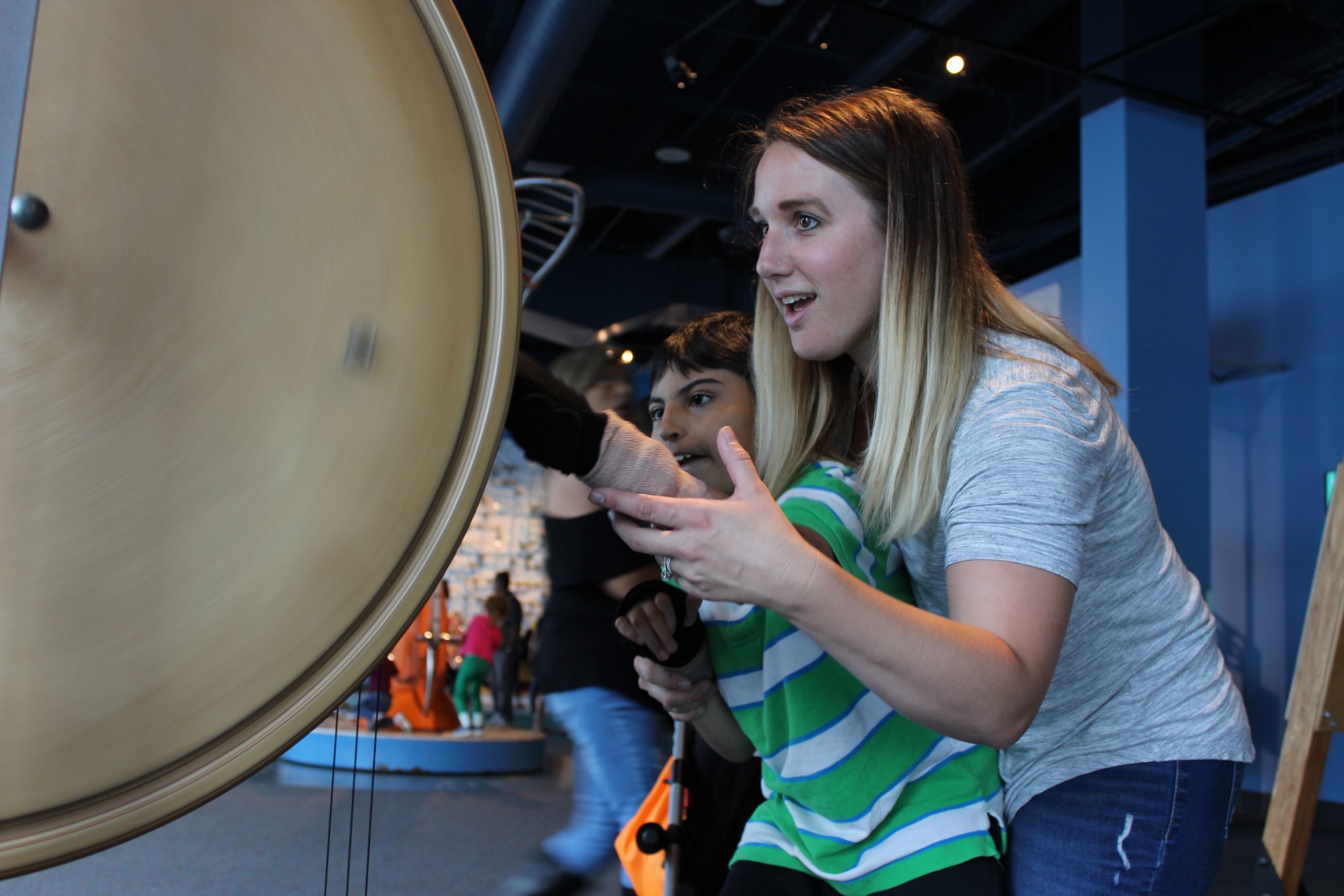 A woman supporting a boy as he stands in front of a large metal disk. They are reaching for the disk.