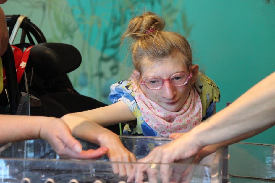 A girl in a wheelchair with her hair in a bun and wearing pink glasses and a pink flowered scarf putting her hand in a container of water with two other adults.