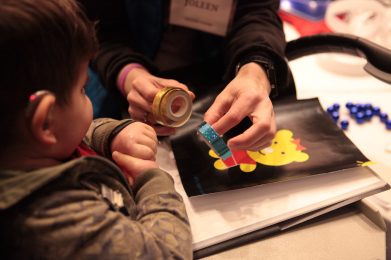 An adult is holding a small roll of yellow ribbon and a small roll of blue ribbon. A young boy is looking at the ribbon.