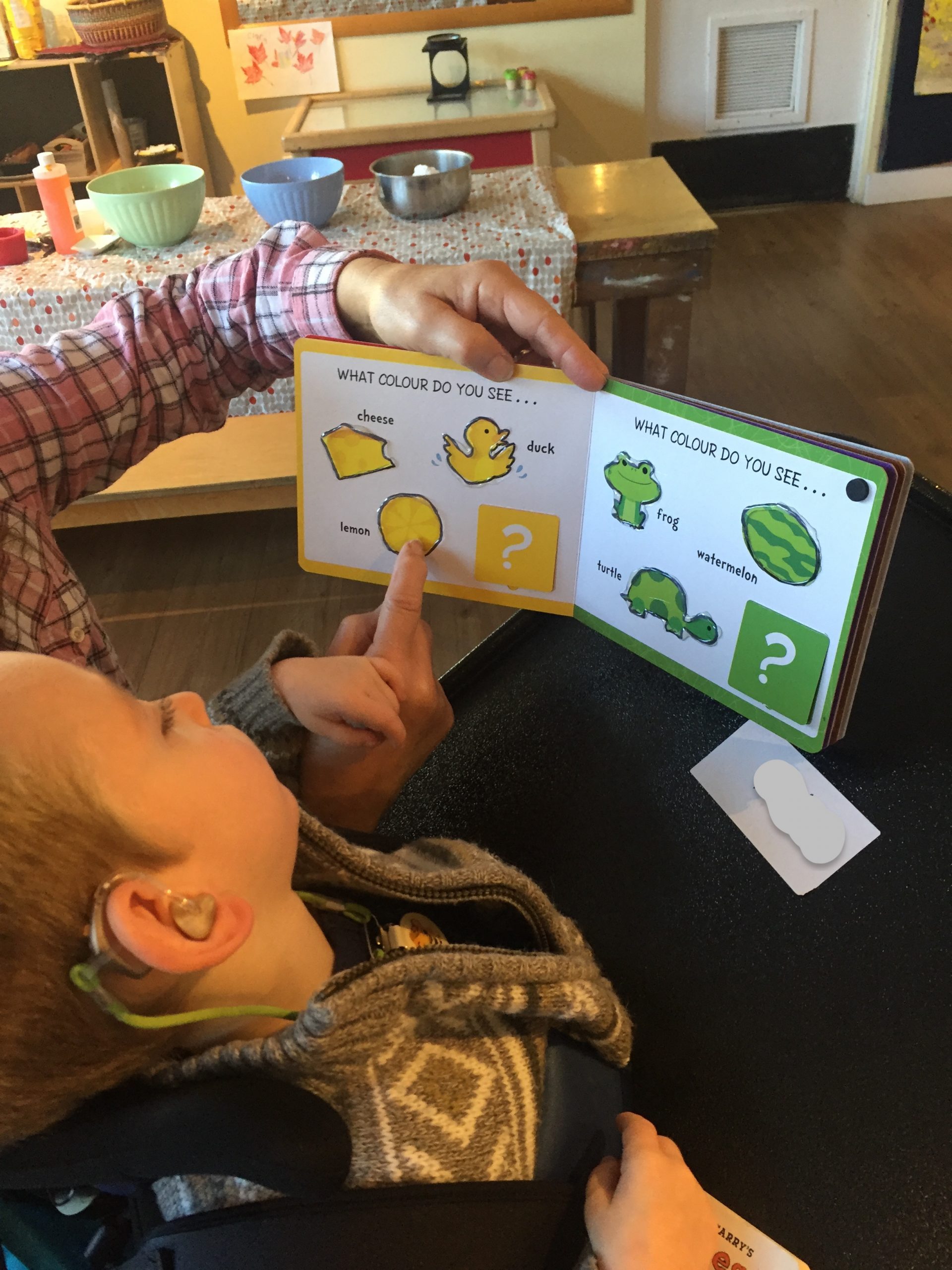 A young boy with hearing aids is in his wheelchair looking at a book with pictures of yellow and green objects. The boy has his hand resting on an adult’s hand, while they point out the objects, using hand-under-hand technique.