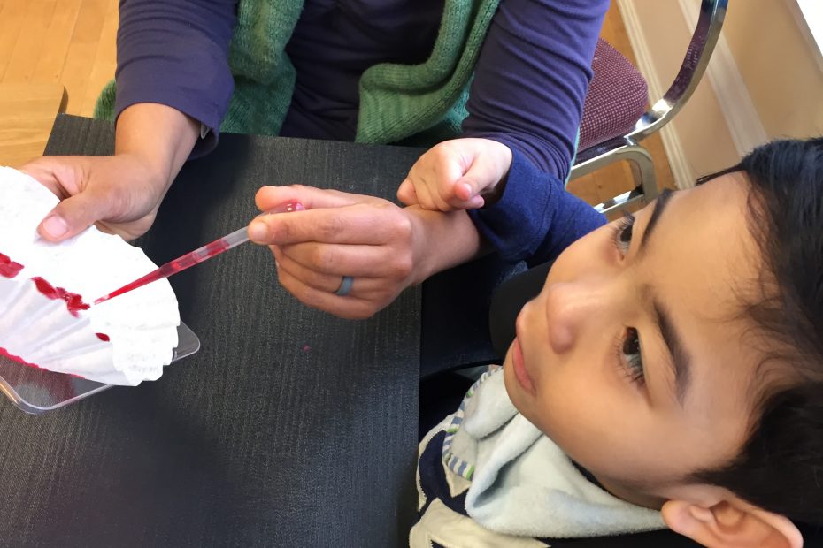 A young boy is watching and resting his hand on an adult’s hand. as the adult paints a coffee filter with a paint brush