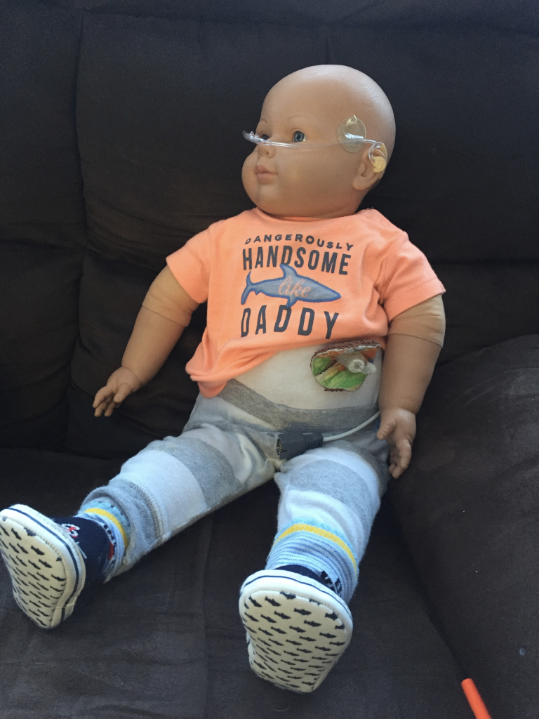 A photo of a male doll wearing an orange t-shirt with ‘Dangerously Handsome Like Daddy’ and a picture of a shark on the front, grey and white striped sweatpants and black sneakers with sharks on the bottom. The doll is wearing a hearing aid and has oxygen tubing in his nose and a g-tube.