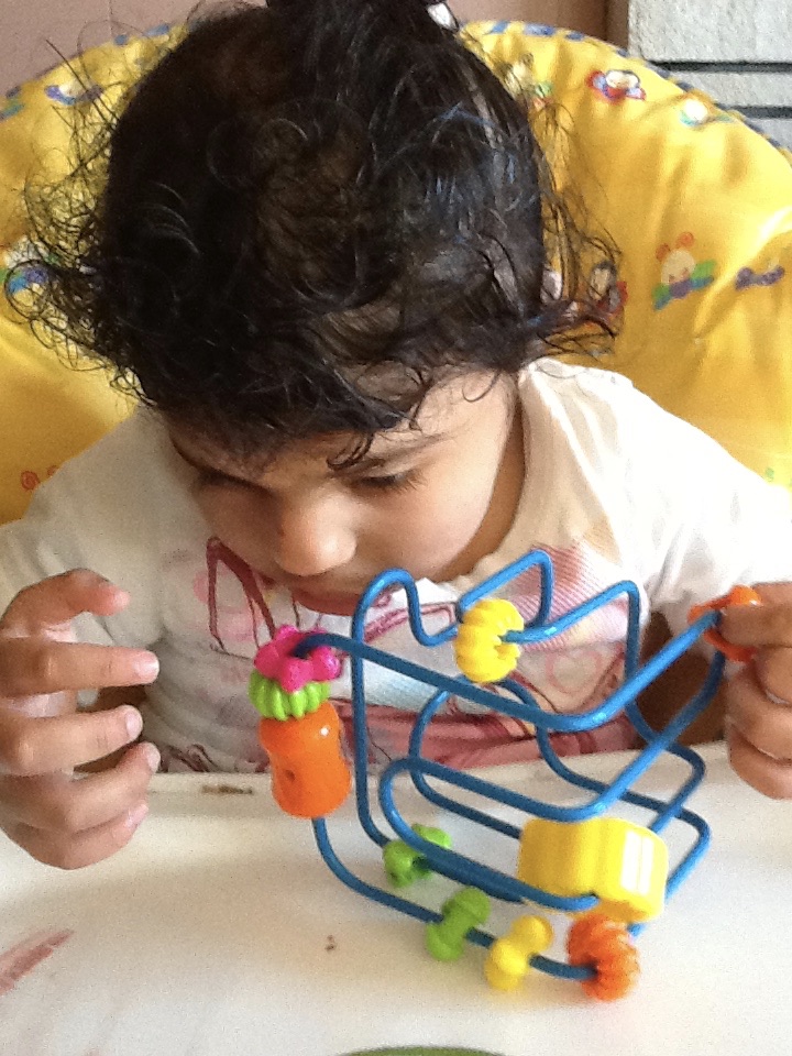 A young girl with dark hair and a white shirt is sitting in her yellow chair playing with a colourful toy.