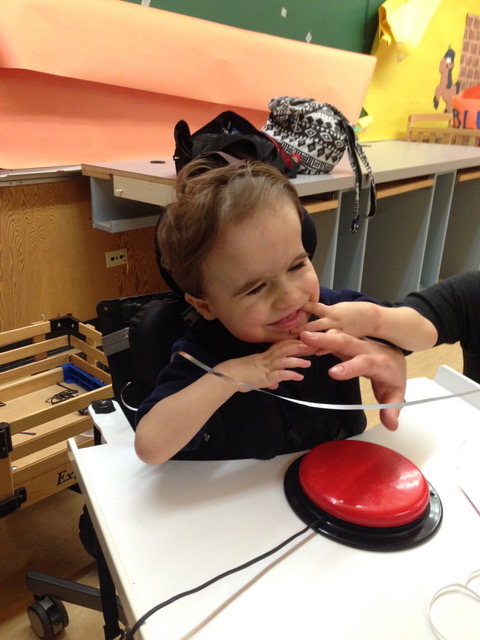 A young boy in a wheelchair wearing a black shirt is smiling while resting his hand on an adult’s hand, using hand-under-hand technique to press a large red switch.