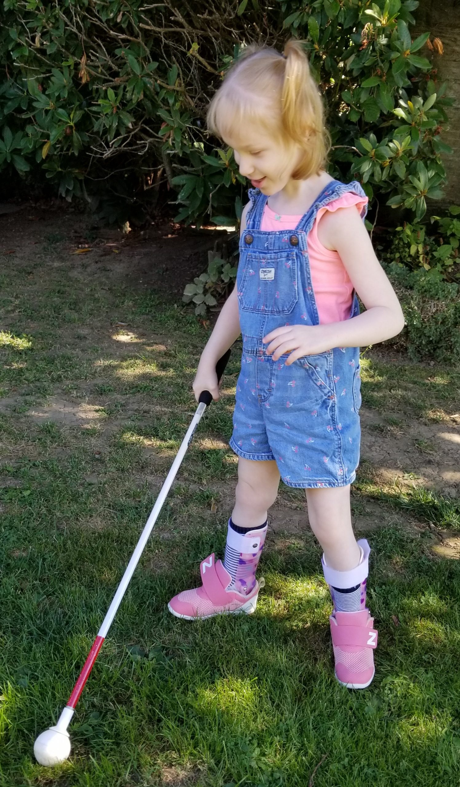 A young girl, smiling, with red hair in a jeans shorts jumpsuit, pink shirt and pink shoes walking on the grass with a white cane.