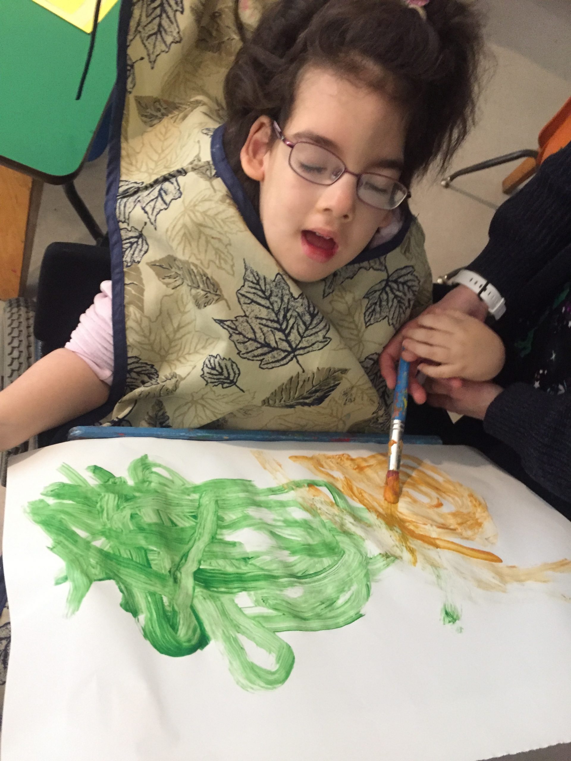 A young girl with dark wavy long hair wearing a bib with sketches of maple leaves on it is painting an orange and green picture. An adult on her left is supporting her left hand as they use hand-under-hand technique to paint with a blue paint brush.